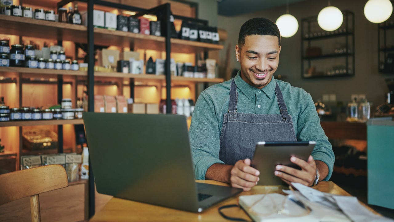 A shop owner wearing an apron smiles as he works on a tablet and laptop in his shop.