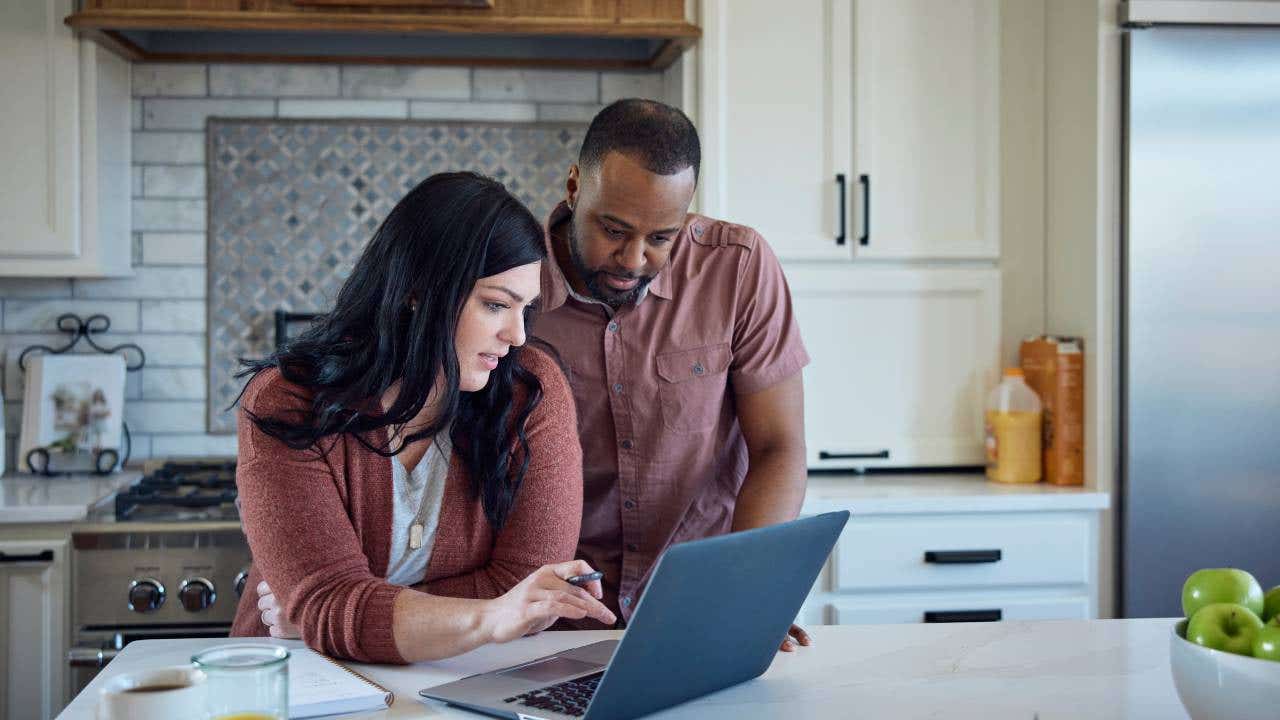 Caucasian woman and African American man sit at kitchen counter with breakfast working with pen, paper and laptop.